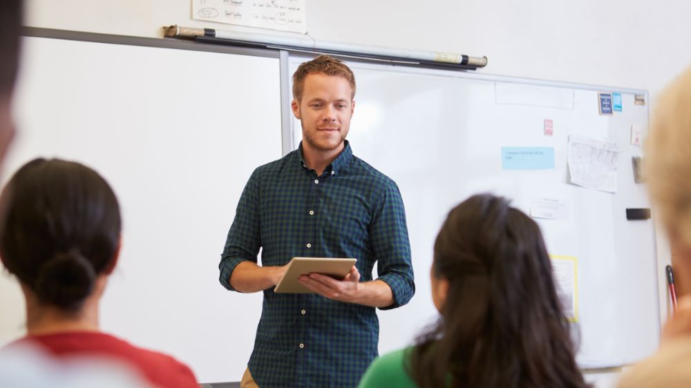 ung mann står foran whiteboard-tavle i klasserom og snakker.