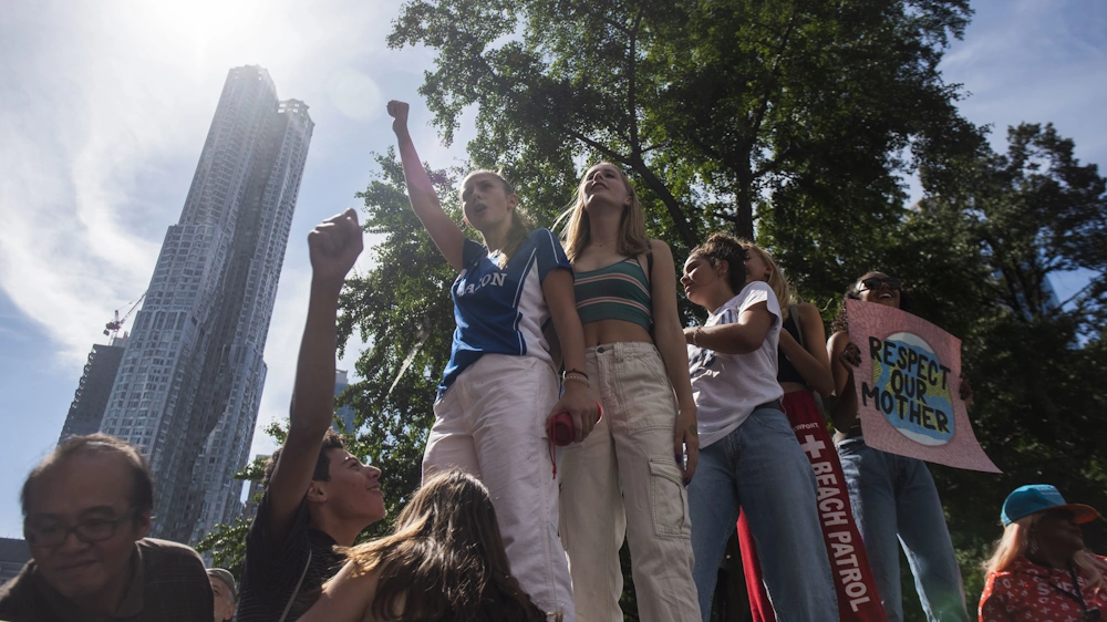 Youth protesting for climate in New York. Five teenage girls in front, hands are raised, paroles held. Tall building behind. Sun shines.