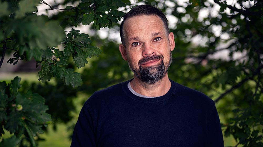 A man in a black sweater with black hair and a black beard stands before an oak wood, smiling.