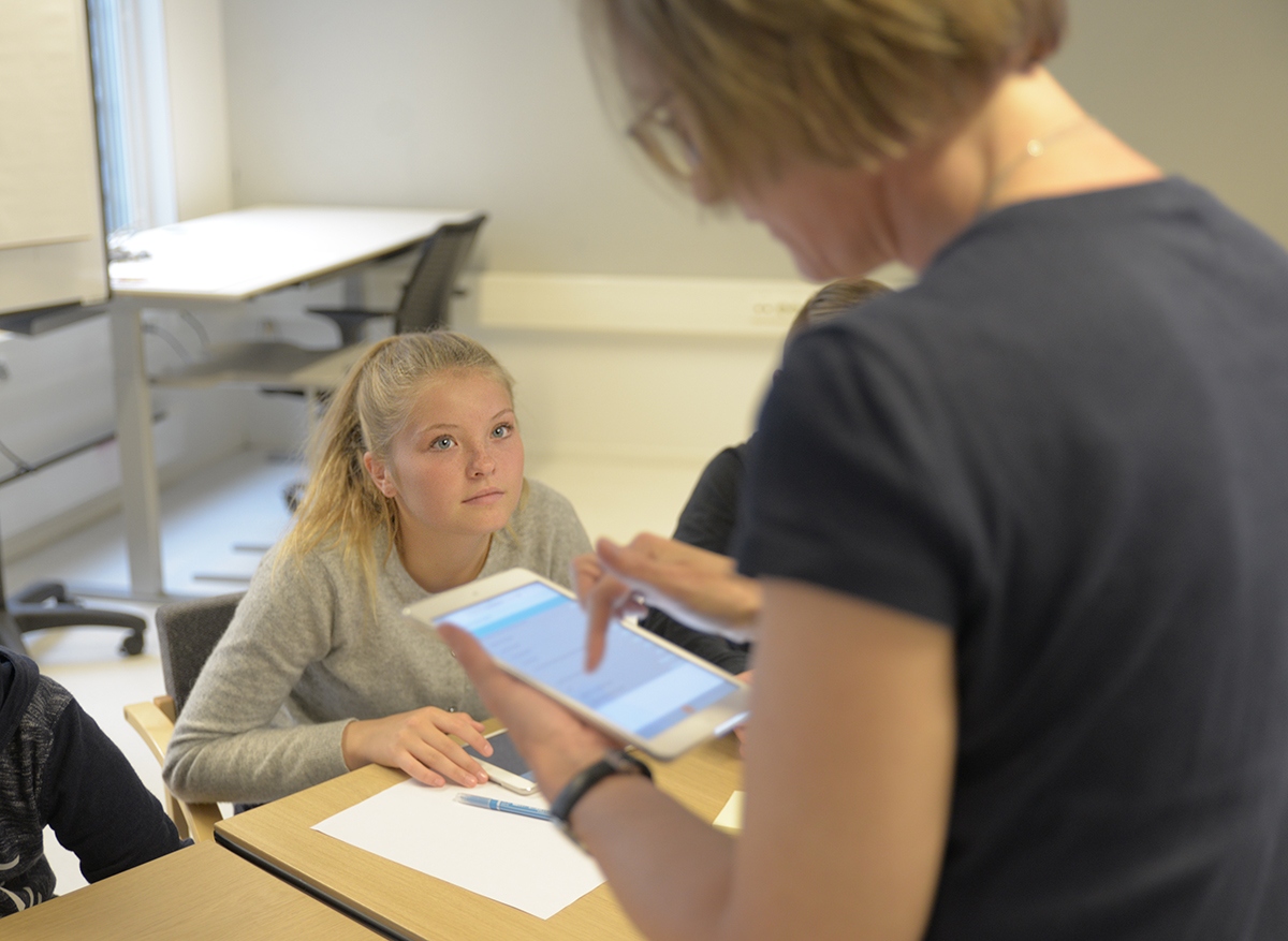 Jente på ungdomstrinnet bak pulten, lærer med nettbrett foran. Fotot