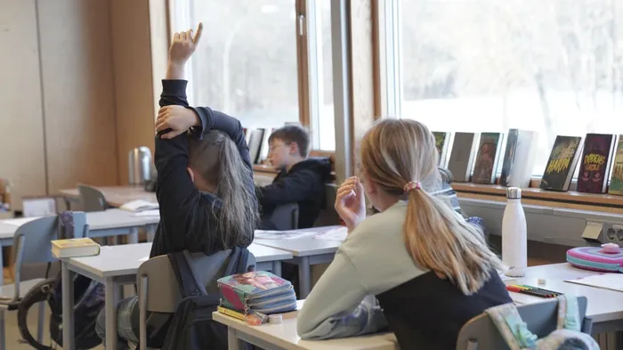 Barn som sitter i et klassrom, bilde er tatt bak i fra, alle ser fremover, ei jente som rekker opp hånda. 