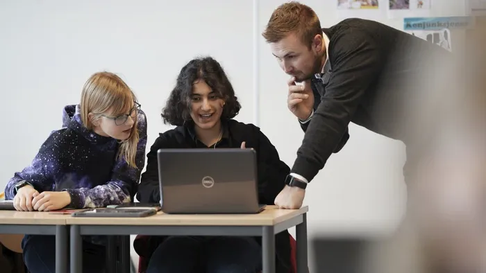 En lærer hjelper to elever i et klasserom, de begge ser på en datamaskin