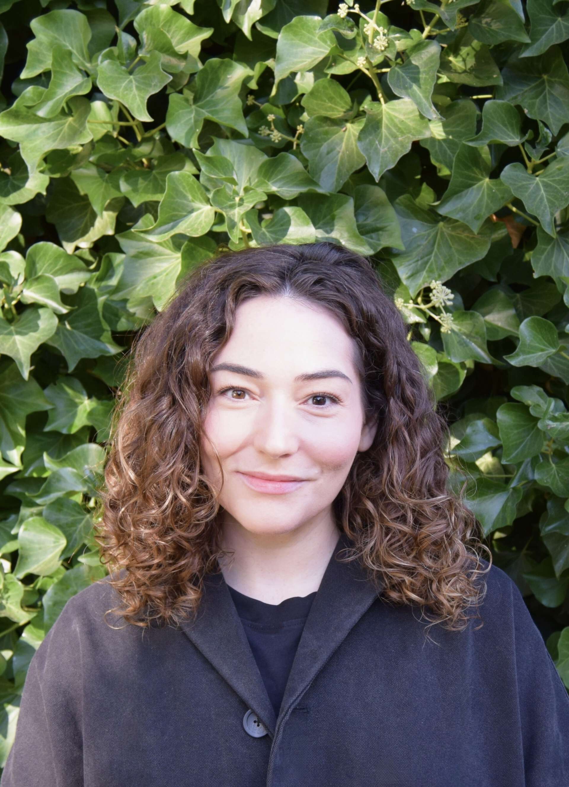 Profile picture of Amalie Fishberg, woman, brown curliy hair. In front of a hedge.