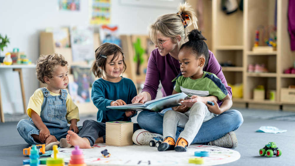 Three children and an adult sitting on a rug in a kindergarten, reading a book together while the adult points at something on the page, with toys scattered on the floor.