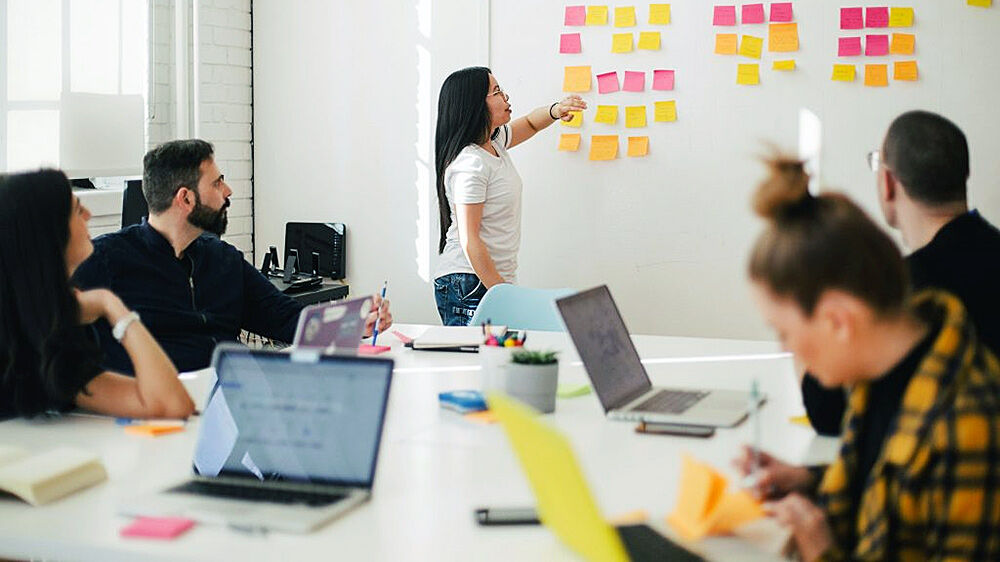 Kollegaer i et møterom med bærbare PC-er, som deltar i et Scrum-møte med Post-it-lapper på veggen.