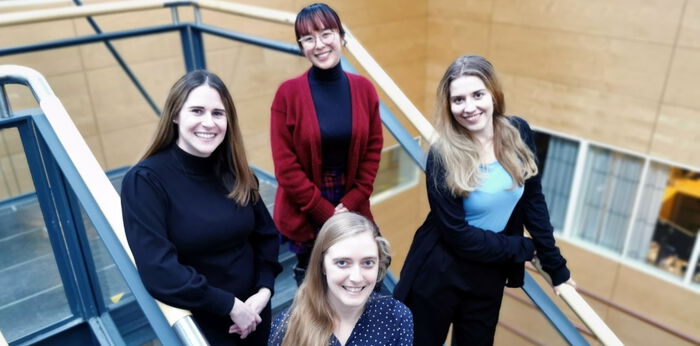 Four female students in their twenties, standing at stairs, smiling.