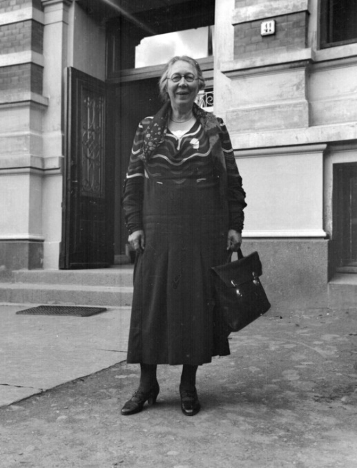 Eldre, svart-hvitt bilde av kvinne foran et bankbygg. Bærer dokumentmappe i hånda og har runde briller og knute i nakken. TAtt i 1933.