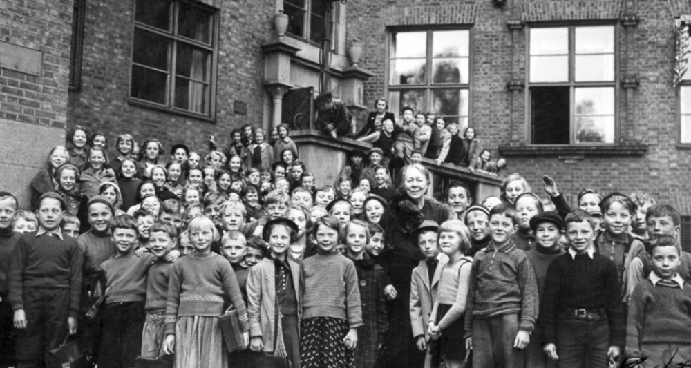Eldre, svart-hvitt bilde av 30-40 barneskoleelever sammen med en kvinnelig lærer, foran et skolebygg. 