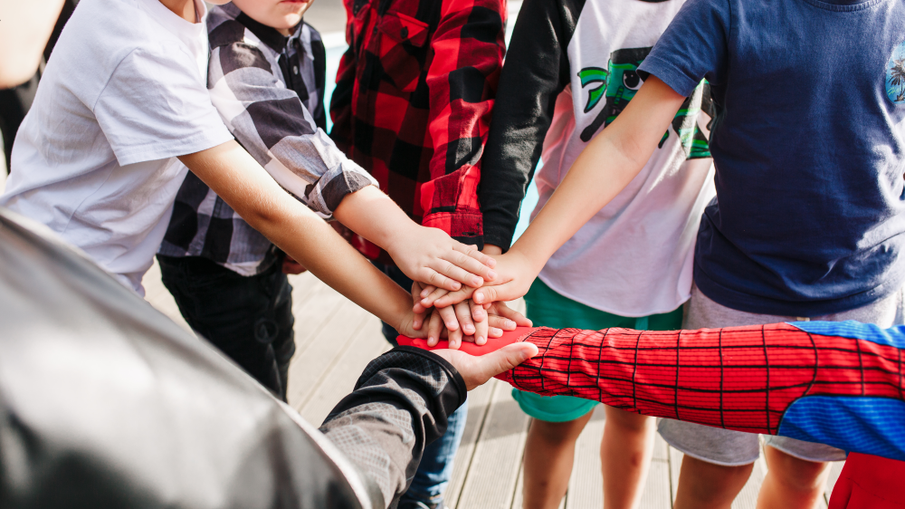 Barneskoleelever står i ring og legger hendene oppå hverandre. En av hendene/armene er utkledd som superhelten Spider-Man med rødt og blått kostyme.