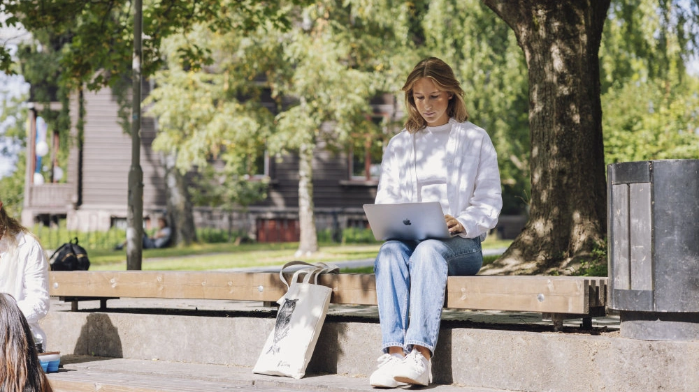 Illustrasjonsfoto. Kvinnelig student sitter med en mac-datamaskin i fanget, på en benk, under et tre på Blindern campus.