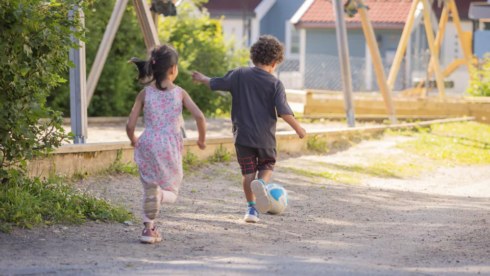 en jente og en gutt springer etter en ball i et utendørs barnehagemiljø.