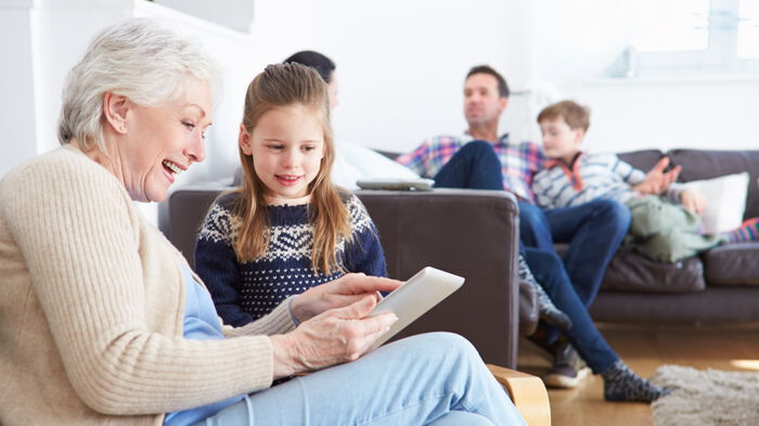 Grandmother and granddaughter sitting together in a living room. Looking at a tablet. Rest of the family sitting in the background.