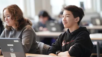 Two teenagers in classroom. Smiling, sitting with laptop, looking at teacher
