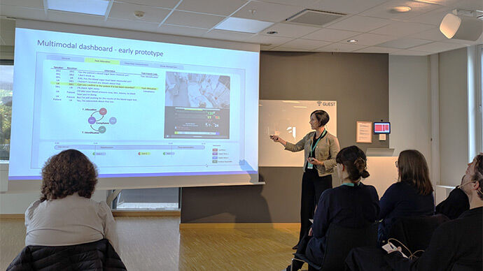 En foredragsholder peker på en PowerPoint-presentasjon mens et publikum følger med i et rom med dempet belysning.