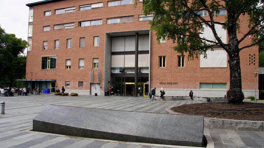 Helga Engs hus på Blindern i Oslo er en mursteinsbygning med brosteinsplass foran hovedinngangen. Plassen har et stort tre til høyre, og det sitter folk på noen av benkene foran bygget.