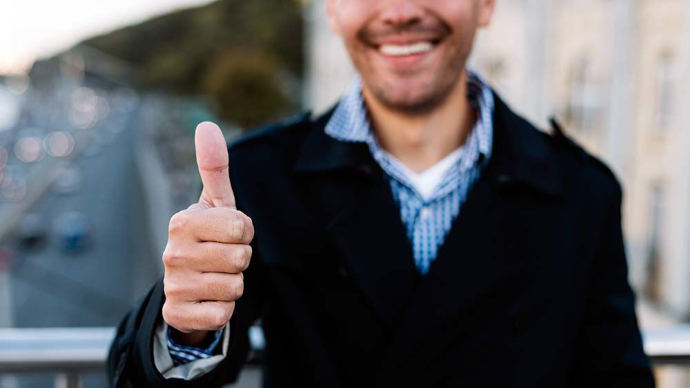 En dresskledd mann står i front og viser tommel opp-tegnet med hånden sin. Han ser direkte inn i kameraet med et smil, og bildet formidler en positiv og selvsikker holdning.