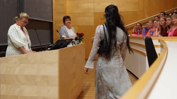 to kvinner står ved et podium i en forelesningssal. En kvinne med ryggen til kamera går mot dem for å få rose. Flere unge studenter sitter i salen.