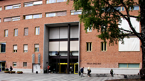Helga Engs hus på Blindern i Oslo er en mursteinsbygning med brosteinsplass foran hovedinngangen. Plassen har et stort tre til høyre, og det sitter folk på noen av benkene foran bygget.