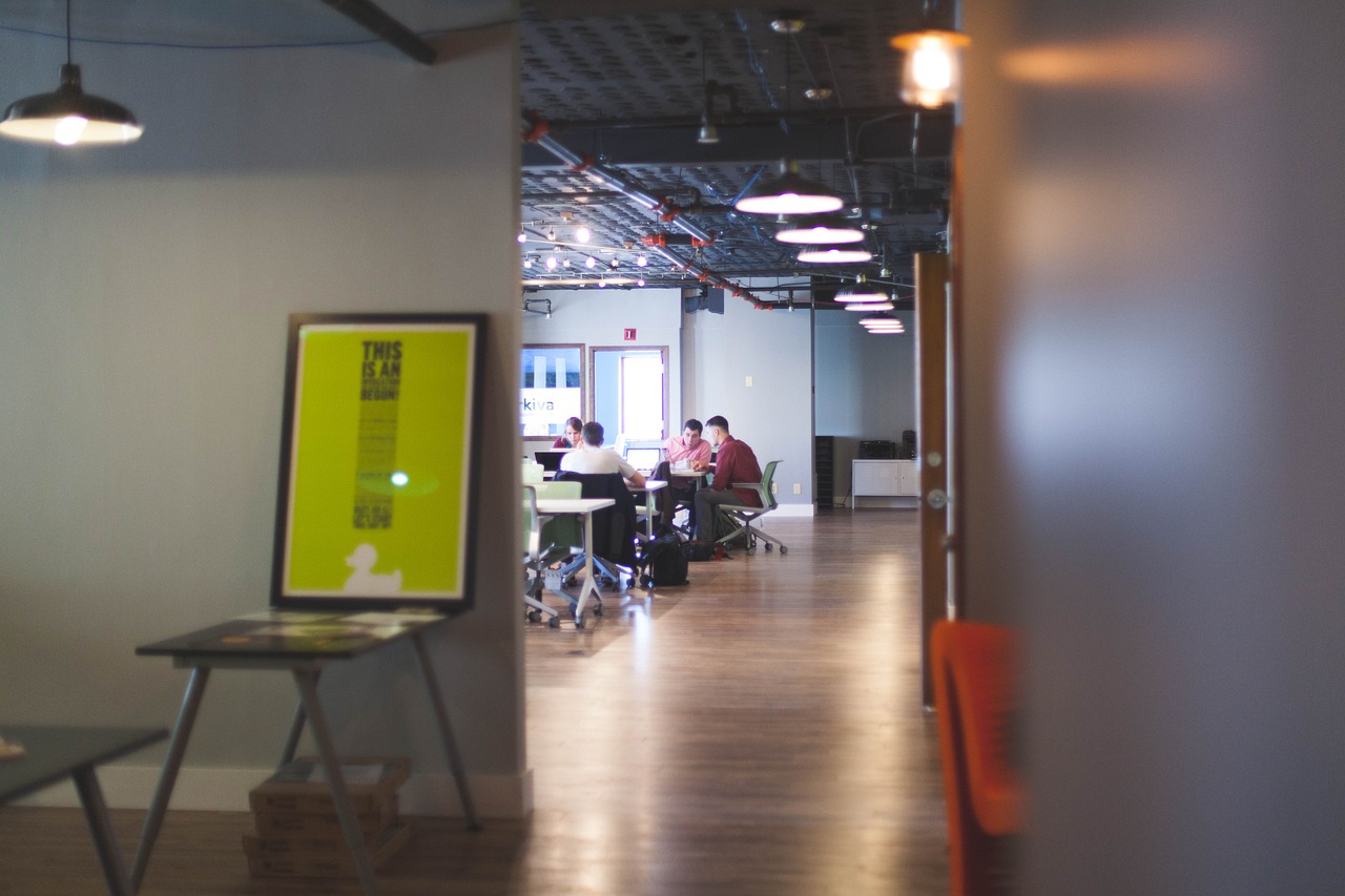 Bildet viser en innendørs setting, muligens et moderne kontor eller et samarbeidsrom. Folk jobber ved et langt bord, opplyst av taklamper. I forgrunnen er det en lys gul plakat på et staffeli med tekst og grafikk.