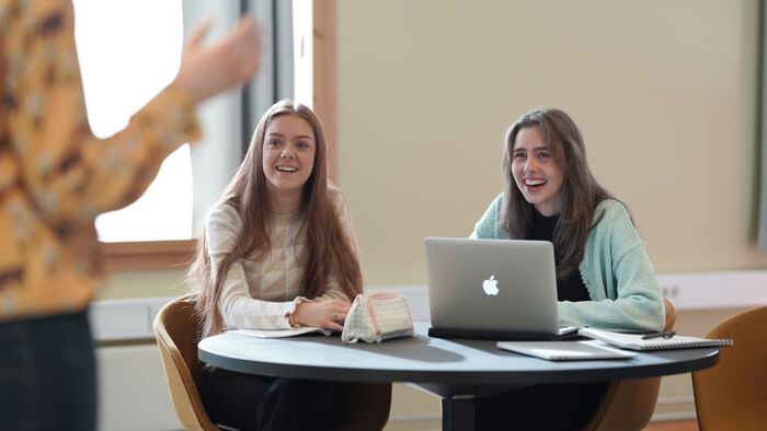 To studenter sitter ved et bord med notatb?ker og en PC. Foran i bildet ser man en tredje person som snakker til studentene.