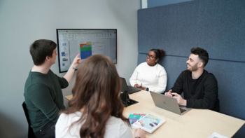 A group of four students sit around a table and share a presentation on a display screen mounted to the wall.